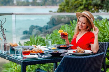 Akşam yemeğinde şapkalı ve elbiseli genç bir kadın tropik bir plajda lüks bir tatil köyünde bir bardak viskiyle. Deniz kenarındaki kafede akşam, okyanus manzaralı..