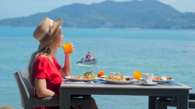 Şapkalı kadın, tropik plajdaki lüks otel otelinde deniz manzaralı kahvaltıdan hoşlanıyor. Sahildeki kafede nefis yemeklerin tadını çıkaran bir kız. Tatil, deniz tatili konsepti.