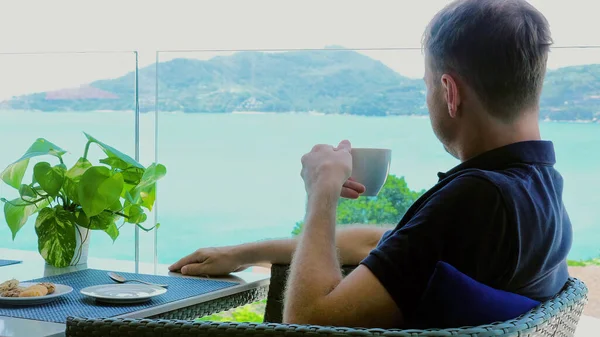 A young man on vacation sits in hotel cafe terrace with view of sea. Male traveler enjoy lunch break, drinking a cup of hot coffee in a luxury restaurant overlooking the ocean.