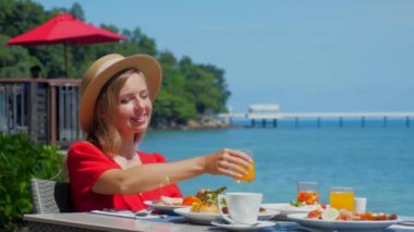 Restoranda tabakları deniz manzaralı yemekle dolu bir masada oturan genç bir kadın. Lüks bir otelde açık büfede kahvaltı yapan bir kız. Seyahat yaşam tarzı. Yaz tatili.