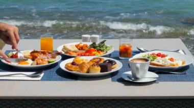 Çeşitli yumurta, sosis, kahve, taze portakal suyu, kruvasan ve deniz manzaralı salata ile servis edilen lezzetli büfe kahvaltısı. Erkekler yemeğin tadını çıkarır, dışarıda yemek yer, cennet gibi okyanus manzarasının tadını çıkarır..