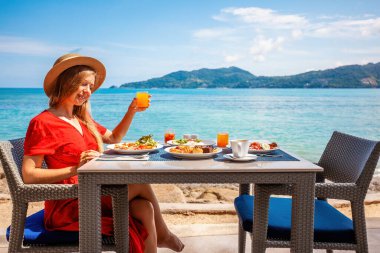 Lüks tatil beldesindeki tatilci kız, taze yiyecek ve tropikal deniz manzaralı sabah kahvaltısının tadını çıkarıyor. Sahilde yemek yiyen mutlu genç bir kadın. Seyahat, tatil, yaz tatili kavramı