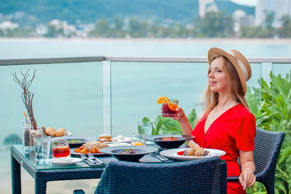 Tatil tatilindeki güzel bir kız alkollü bir kokteyl ile lezzetli bir akşam yemeği yiyor. Tropikal bir plajda açık hava restoranı, deniz manzarasının tadını çıkarıyor..
