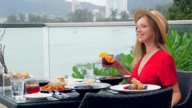 Akşam yemeğinde şapkalı ve elbiseli genç bir kadın tropik bir plajda lüks bir tatil köyünde bir bardak viskiyle. Deniz kenarındaki kafede akşam, okyanus manzaralı..