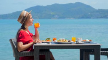 Tatil tatilindeki blogcu kadın, tropik plaja bakan lüks bir otel restoranında kahvaltıdan zevk alıyor. Etkilenen kız, akşam yemeği yerken turkuaz okyanusun çarpıcı manzarasının tadını çıkarıyor..