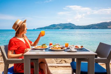 Kadınlar tropikal plajdaki lüks otel otelinde deniz manzaralı kahvaltıdan hoşlanır. Sahildeki kafede lezzetli yemeklerin tadını çıkaran güzel bir kız. Arka planda jet ski var. Tatil, deniz tatili konsepti..