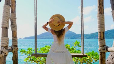 Hasır şapkalı genç kadın salıncaktan okyanus manzarasını seviyor. Deniz kıyısındaki bir tatil beldesinde dinlenme konsepti. Tasasız ve mutlu, yazın özgürlüğü ve doğanın güzelliği..