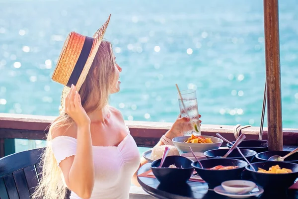 年轻女子在一家豪华酒店度假胜地享用美味的早餐 女孩坐在餐桌前 喝着一杯水 望着外面的大海 头戴草帽的女性享受暑假 图库图片