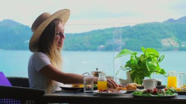 Deniz manzaralı kafe masasında oturan gülümseyen kadın, taze yemeğin tadını çıkarıyor, lüks otellerde açık büfe kahvaltıda hamur işi yiyor. Yaz tatilinde Tayland, Asya 'da Phuket Adası' nda otel kahvaltısı..