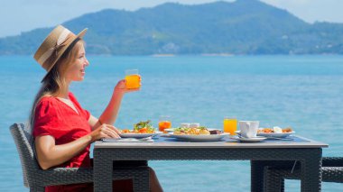 Tatil tatilindeki blogcu kadın, tropik plaja bakan lüks bir otel restoranında kahvaltıdan zevk alıyor. Etkilenen kız, akşam yemeği yerken turkuaz okyanusun çarpıcı manzarasının tadını çıkarıyor..