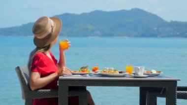 Lüks tatil beldesindeki tatilci kız, taze yiyecek ve tropikal deniz manzaralı sabah kahvaltısının tadını çıkarıyor. Sahilde yemek yiyen mutlu genç bir kadın. Seyahat, tatil, yaz tatili kavramı