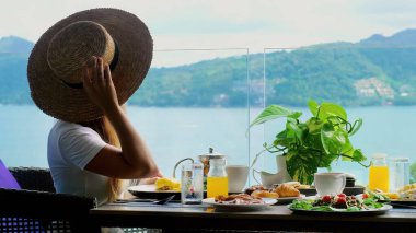 Deniz kenarındaki kafede şapkalı bir kadın kahvaltı büfesinin ya da akşam yemeğinin tadını çıkarıyor. Yaz tatilinde tropik tatil beldesinde otel kahvaltısı. Phuket Adası, Tayland, Asya.