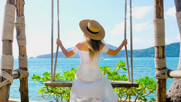 Salıncakta sallanan, denize bakan hasır şapkalı bir kadının arka plan görüntüsü. Güzel bir tatil yeri, tatil kaçamağı, tropik bir cennet. Ada tatili.