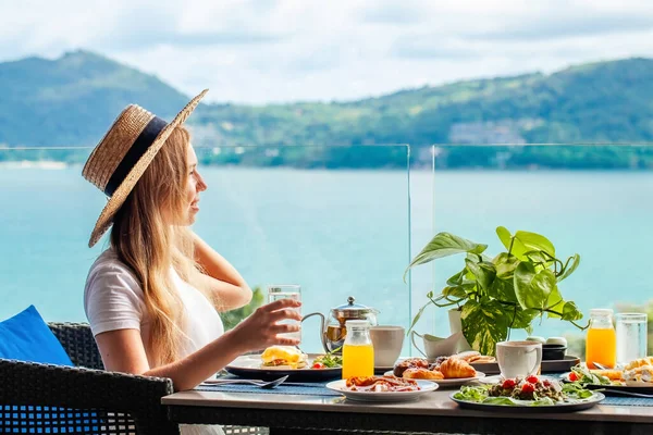 微笑的女人坐在咖啡馆的桌子边 一边欣赏大海 一边喝着一杯橙汁 在旅馆吃早餐 热带度假胜地的暑假 普吉岛 旅游广告 早餐自助餐 免版税图库图片