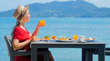 Lüks bir otelde sakin bir sabah. Bir kadın okyanus manzaralı kahvaltısını sever. Tropikal plajın huzurunu ve doğal güzelliğini kucaklar..