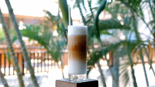 Coffee cold iced drink with ice and milk in outdoor cafe with palm trees on background. Concept of refreshing tropical beverage in a serene cafe setting.