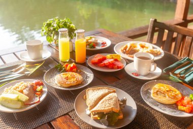 Huzurlu bir göl kenarındaki Tayland tatil köyünde lezzetli yemekleri olan sağlıklı bir kahvaltı masası. Taze meyve, krep ve tropikal tatlar. Kavram: Tayland tatili.