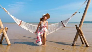 Deniz sahilindeki hamakta dikilen ve okyanus manzaralı sahilde yürüyen mutlu kadın. Tayland, Phuket Adası 'nda tatil. Ağır çekim. Neşeli sahil tatili ve boş zaman kavramı.