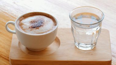 Kafe ahşap masada sıcak kapuçino ve kaynar su. Masa üstünde beyaz bardakta köpüklü latte. Kahve ve sabah. Kremalı sıcak kapuçino.