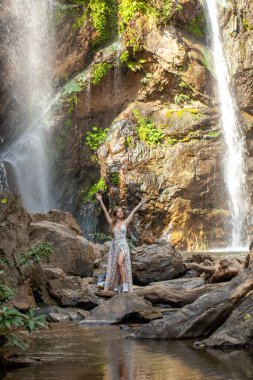 Kolları şelaleden yükselen bir elbise giymiş bir kadın. Bereketli yeşil orman cennetinin manzaralı güzelliğini keşfedin. Doğa ve canlı yeşillik, tropikal bir yer..