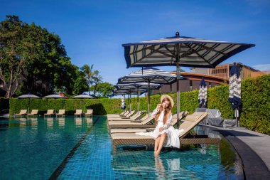 Sakin yaz sahnesi. Beyaz elbiseli bir kadın, modern Tayland villasında havuz başında dinleniyor. Cennetin tropik güzelliği, yemyeşil manzara ve masmavi sular, mükemmel tatil için nihai rahatlama..