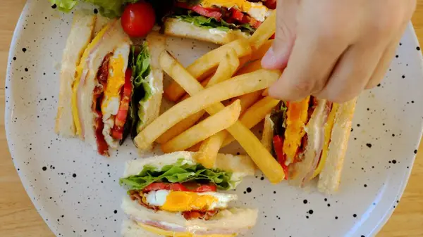 Man hand take a french fries from plate with crunchy and delicious club sandwich with assorted meats, cheese, lettuce, and tomato on toasted bread in cafe. Top view.