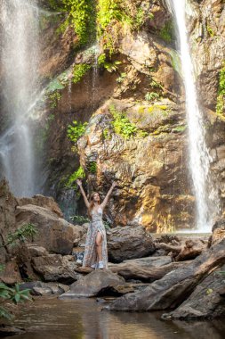 Tropik şelalenin yanında uçan elbiseli bir kadın. Doğa, su ve seyahat, Tayland 'daki yemyeşil orman cenneti manzarasıyla birleşir. Bu ferahlatıcı vahanın güzelliğini keşfedin..