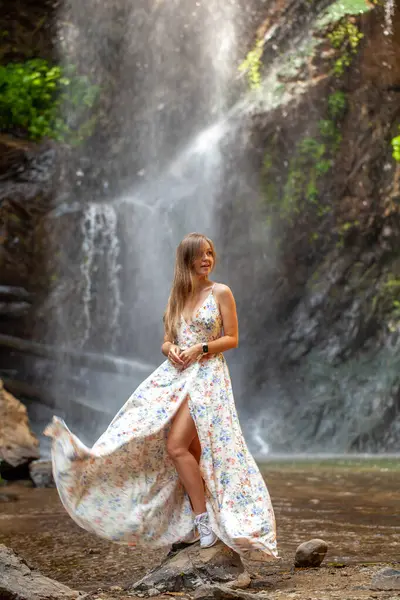 Scenic view of a woman in a flowing dress by a lush forest waterfall. Serenity amidst the natural beauty of Asia.