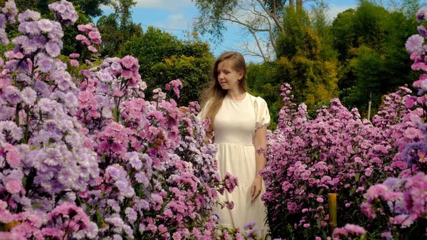 Young woman in flowing white dress walking through field of vibrant violet wildflowers in spring.
