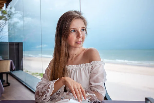 Happy diner in light off-shoulder blouse, engaging in meal, transparent blue ocean backdrop, serene dining experience. Coastal dining and serene vacation