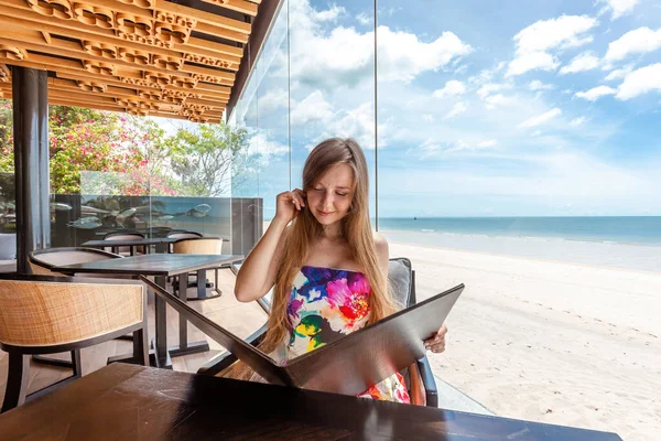Woman in colorful attire at seaside cafe table, engaging with menu, holiday dining with scenic ocean view. Seaside dining and vacation relaxation