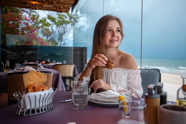 Smiling young woman, casual elegant attire, enjoys bread at sea-view restaurant, clear skies and calm ocean horizon. Culinary experiences and seaside enjoyment