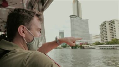 Yüzü maskeli bir adam tekneden şehrin siluetini gösteriyor. Şehir keşfi ve seyahat.