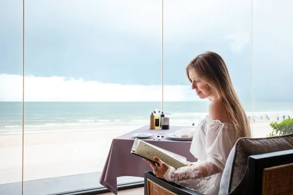 Young adult enjoying menu selection, seaside fine dining, panoramic ocean backdrop, relaxed beach holiday, elegant table arrangement.