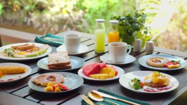 Açık veranda masasında taze meyve, içecek ve ana yemekle birlikte çeşitli besleyici kahvaltı seçenekleri sunuluyor. Sağlıklı yemek ve yaşam tarzı.