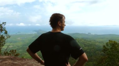 Man standing on mountain peak overlooking scenic landscape with lush greenery and distant ocean. Travel and exploration.