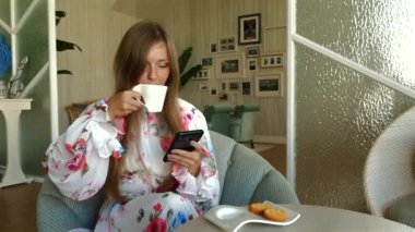Kadın sabah kahvesinden zevk alıyor ve rahat oturma odasında akıllı telefon kullanıyor. Modern yaşam tarzı ve rahatlama.