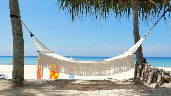 White hammock tied between palm trees on tropical beach with turquoise sea in background. Vacation and travel relaxation.