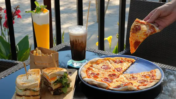 Dining experience with variety of fast food items including fresh pizza slice being picked up, sandwich with fries, and assorted beverages on sunny patio. Casual dining and fast food.