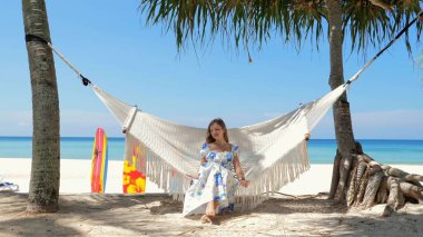 Huzurlu tropikal plaj tatili yapan bir kadın, arka planda sörf tahtaları olan palmiye ağaçları arasındaki hamakta dinleniyor. Boş zaman yolculuğu ve rahatlama..