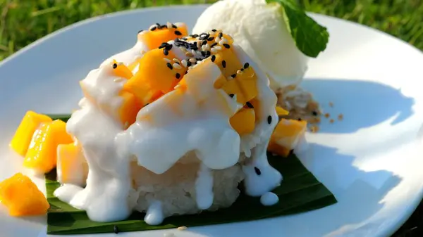 Gourmet tropical dessert of sticky rice, mango, and coconut milk on white plate garnished with sesame seeds and mint, served with ice cream. Presentation and plating of exotic sweets.