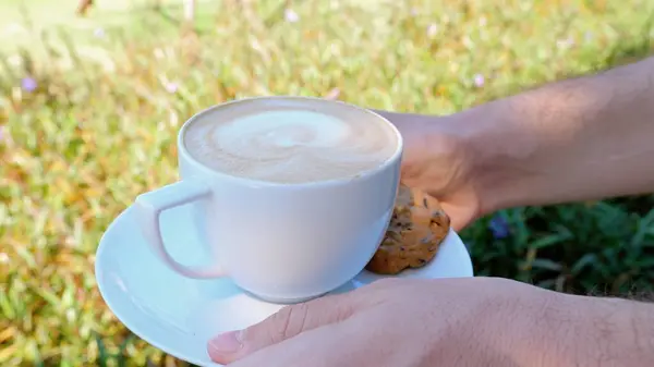 The person is holding a cup of coffee and a cookie on a saucer with their thumb and finger. Coffee on the grass background, relaxing moment of sharing food.