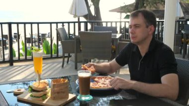 Sahildeki restoranda pizza ve sandviçle yemek yiyen bir adam. Arka planda tropik okyanus manzarası olan. Rahatlama ve tatil yemeği deneyimi.