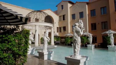 Modern mimari ve yemyeşil alanlarda berrak mavi gökyüzü altında durgun su havuzunu çevreleyen klasik Greko-Romen heykelleriyle açık avluda. Seyahat, mimari ve sanat için ideal.
