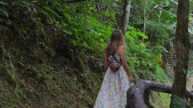 Çiçekli elbiseler giyen genç bir kadın gür yeşil orman yolunda yürüyor. Ağaçlar ve yapraklar sık, sırt çantası taşıyor. Doğa ve macera.