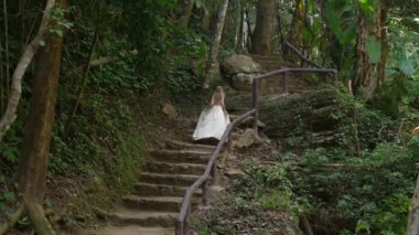 Beyaz elbiseli kadın yeşil yemyeşil orman ve tropikal bitki örtüsüyle çevrili taş basamaklarda yürüyor. Yol ahşap korkuluklarla kaplı, ve sahne keşif ve doğa duygusunu çağrıştırıyor..