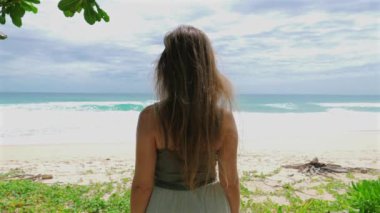 Kumlu sahilde durup okyanus dalgalarına bakan uzun saçlı bir kadın. Gökyüzü parçalı bulutlu ve yemyeşil plaj alanını çevrelemektedir. Rahatlamak ve günlük rutinden kaçmak.
