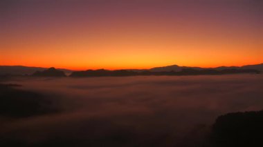 Şafak vakti sislerle kaplı dağların panoramik manzarasıyla nefes kesici bir gün doğumu manzarası. Rengi koyu turuncudan mor renge dönüşen, dingin ve huzurlu bir gökyüzü.