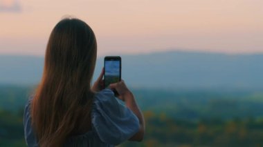 Gün batımında akıllı telefonuyla nefes kesici manzarayı yakalayan uzun saçlı kadının arka plan görüntüsü. Doğa fotoğrafçılığı meraklısı canlı renklerle manzaranın tadını çıkarıyor. Teknoloji ve seyahat