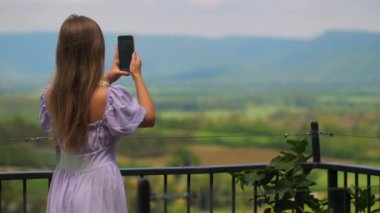 Lavanta elbiseli kadın balkonda dikiliyor, akıllı telefonuyla manzaranın fotoğrafını çekiyor..